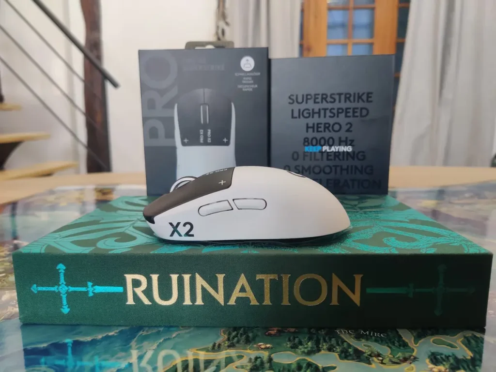 Logitech G Pro X2 Superstrike alb vazut din lateral, cu butoanele laterale vizibile, asezat pe cartea Ruination, cu specificatiile HERO 2, 8000Hz si 0 Filtering vizibile pe ambalaj in fundal